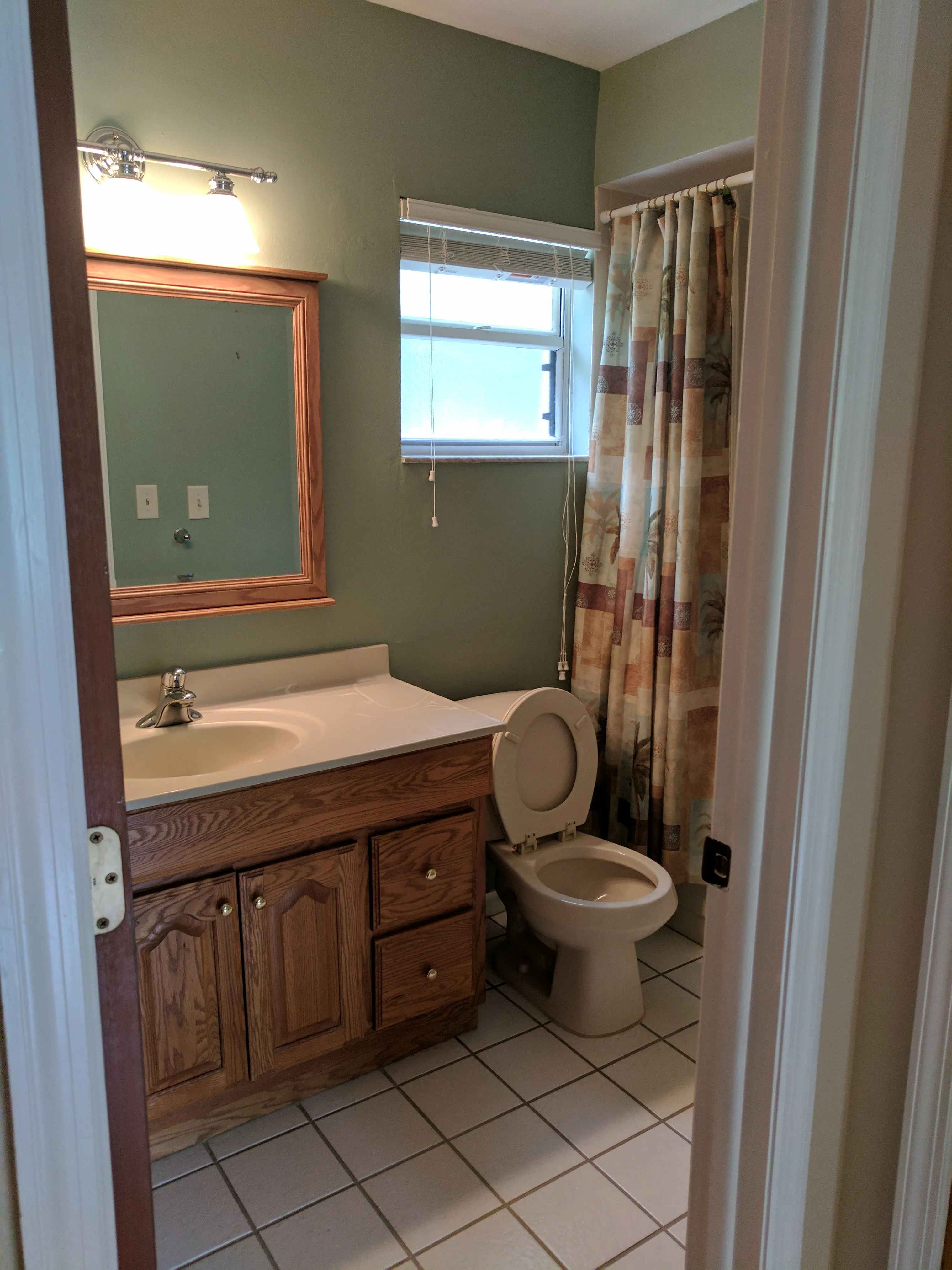 Before: Bathroom refreshed from a dated oak-and-sage look to a bright coastal style — white shaker vanity, quartz countertop, mother-of-pearl tile mirror frame and backsplash, sea-glass linear shower tile behind a glass panel, marble hexagon floor tile, and fresh neutral paint