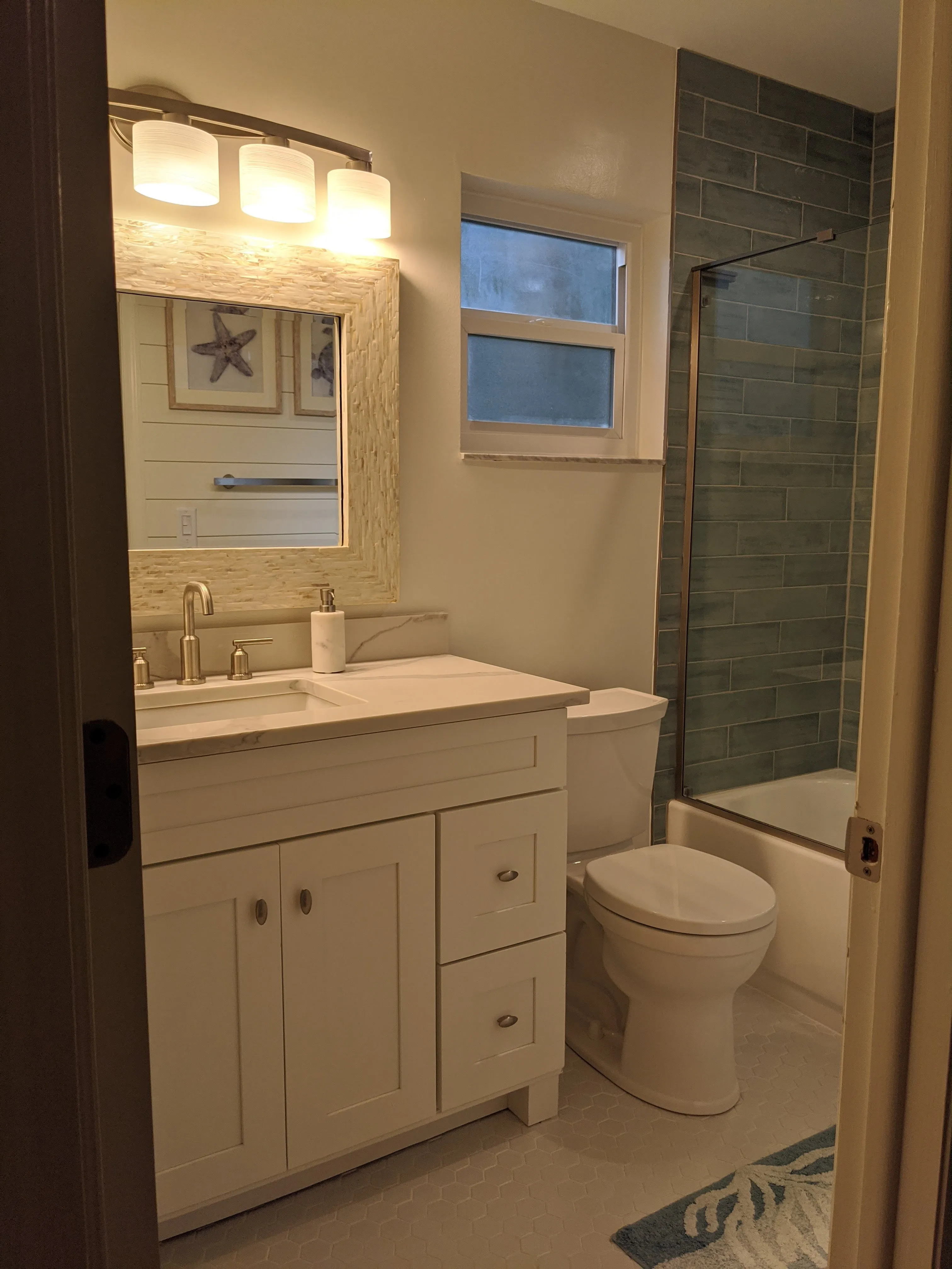 After: Bathroom refreshed from a dated oak-and-sage look to a bright coastal style — white shaker vanity, quartz countertop, mother-of-pearl tile mirror frame and backsplash, sea-glass linear shower tile behind a glass panel, marble hexagon floor tile, and fresh neutral paint